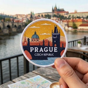 May include: A circular embroidered patch featuring a stylized cityscape of Prague, Czech Republic. The design includes a dark blue bridge tower, a domed building, and a yellow sky with white clouds. The text PRAGUE CZECH REPUBLIC is stitched in white.