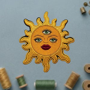 May include: Embroidered patch featuring a stylized sun with three eyes, a red mouth, and yellow rays. The patch has a detailed design with green eyes and is likely for clothing or accessories.