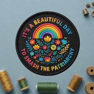 May include: Embroidered black patch with the text "It's a beautiful day to smash the patriarchy." The patch features a rainbow and colorful floral design. Sewing supplies, including spools of thread and a thimble, are in the background.
