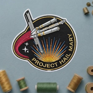 May include: Embroidered patch with a black background featuring a spaceship design, the text "PROJECT HAIL MARY", and a red and gold border. The patch includes a stylized sunburst design.