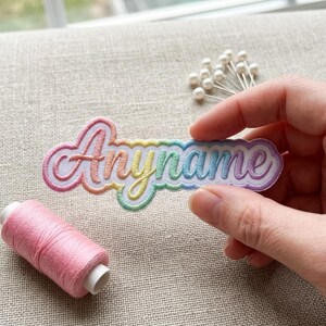 May include: An embroidered patch featuring the word "Anyname" in a cursive script. The lettering displays a gradient of rainbow colors, including pink, orange, yellow, green, blue, and purple, with a white outline and a scalloped border.