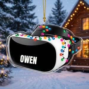 May include: A clear acrylic ornament shaped like a virtual reality headset, featuring a black front panel with the name OWEN printed in white capital letters. The headset is wrapped in a string of colorful, glowing Christmas lights and hangs from a gold cord.