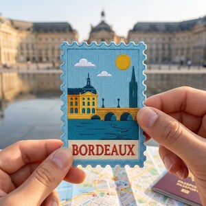 May include: A rectangular embroidered patch shaped like a postage stamp with a light blue border. The design features a yellow building, a stone bridge over blue water, a yellow sun, and white clouds. The word BORDEAUX is stitched in red letters.