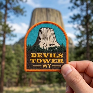 May include: A semi-circular embroidered patch featuring a depiction of Devils Tower in Wyoming. The design includes a light-colored rock formation against a teal sky, dark green pine trees, and the yellow text "DEVILS TOWER WY" on a brown base.