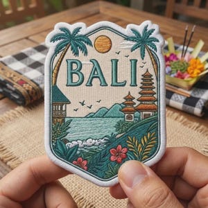 May include: A shield-shaped embroidered patch featuring the text BALI in teal letters. The design depicts a tropical landscape with palm trees, a tiered temple, mountains, a sun, birds, and red flowers against a cream-colored background.