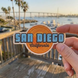 May include: A circular embroidered patch featuring a stylized orange and yellow sunburst background. The text SAN DIEGO is stitched in light blue with dark blue outlines, and the word California is embroidered in dark brown cursive script.