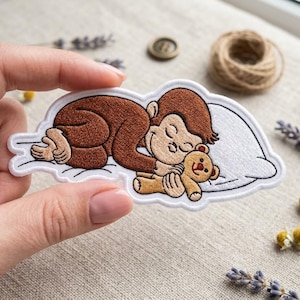 May include: An embroidered patch featuring a brown monkey sleeping on a white pillow while hugging a small tan teddy bear. The patch has a white border and is shaped like a cloud, showcasing detailed stitching of the monkey's fur and facial features.