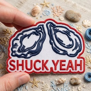 May include: An embroidered patch featuring two dark blue oyster shells outlined in red on a white background. The text SHUCK YEAH is embroidered in bold red capital letters at the bottom of the patch. The design has a red border.