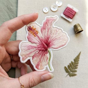May include: A fabric patch featuring an embroidered hibiscus flower in shades of light pink, dark pink, and cream with a yellow stamen. The flower has a white stitched border and a green stem, showcasing detailed thread work and texture.