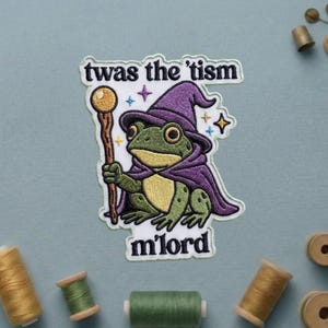 May include: Embroidered patch featuring a green frog dressed as a wizard, wearing a purple hat and cape, holding a staff. The text "twas the 'tism m'lord" is above and below the frog. The patch is on a light blue background.