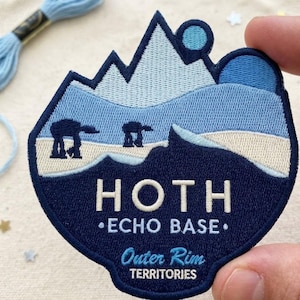 May include: An embroidered patch featuring a snowy mountain landscape in shades of blue and white. The design includes two dark blue AT-AT walkers and the text HOTH ECHO BASE Outer Rim TERRITORIES in white lettering against a dark blue background.