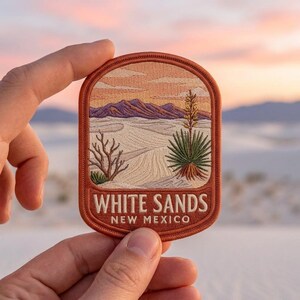 May include: An embroidered patch with a rounded top edge featuring a desert landscape with white sand dunes, purple mountains, a green yucca plant, and a bare shrub under a peach sky. The text WHITE SANDS NEW MEXICO is stitched in white.