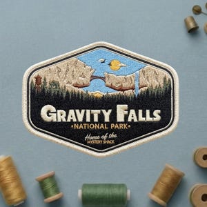 May include: Embroidered patch featuring a landscape scene with the text "Gravity Falls National Park." The patch has a black border and a cream-colored outline. The design includes a bridge, waterfall, and trees, with the words "Home of the Mystery Shack."