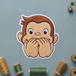May include: An embroidered patch featuring a cartoon monkey with brown fur and a tan face, covering its mouth with both hands. The design is outlined in white thread, showcasing detailed stitching that defines the facial features and fur texture.