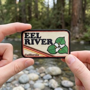 May include: A rectangular embroidered patch with a cream-colored background, featuring the black text EEL RIVER and California. The design includes a white trillium flower with green leaves and three diagonal stripes in orange, yellow, and brown.