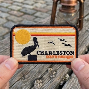 May include: A rectangular embroidered patch featuring a black pelican silhouette perched on a post, a yellow sun, and three flying birds against a cream background. The text reads CHARLESTON SOUTH CAROLINA in black and orange, framed by orange stripes.