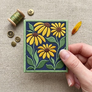 May include: A rectangular embroidered patch featuring three yellow flowers with brown centers and green leaves on a dark blue background, bordered by a green stitched edge. The patch is displayed alongside a spool of green thread and buttons.