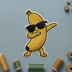 May include: An embroidered patch featuring a yellow banana character wearing black sunglasses and performing a dabbing pose. The banana has a curved body, small arms and legs, and a black tip at the bottom, outlined with a white border.