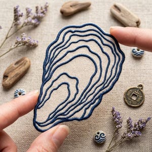 May include: An embroidered patch featuring a dark blue, wavy, topographical-style line pattern on a light beige fabric base. The patch has an irregular, organic shape resembling an oyster shell with concentric, flowing lines throughout.