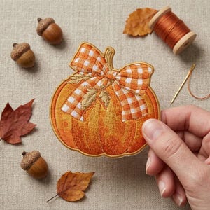 May include: An embroidered pumpkin patch featuring a vibrant orange pumpkin with a white and orange gingham bow. The patch is surrounded by three brown acorns, two dried autumn leaves, a wooden spool of orange thread, and a gold sewing needle.