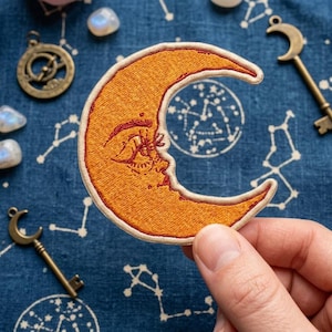 May include: An embroidered crescent moon patch featuring a warm orange hue with a detailed face design, outlined in a light cream border. The moon has closed eyes and subtle facial features stitched in a darker orange thread.