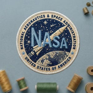 May include: Embroidered NASA patch featuring a rocket, Earth, and stars on a navy blue background. The patch is circular with the text "NATIONAL AERONAUTICS & SPACE ADMINISTRATION" and "UNITED STATES OF AMERICA". The NASA logo is in light blue.