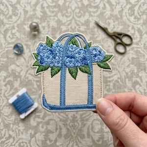 May include: An embroidered patch featuring a beige tote bag with blue handles and trim, overflowing with vibrant blue hydrangea flowers and green leaves. The design is rendered in detailed thread work with a clean, defined border.