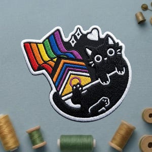 May include: Embroidered patch featuring a black cat with large white eyes and a rainbow flag design. The cat is holding a ring, and the patch has a white border. The patch is on a light blue background with spools of thread.