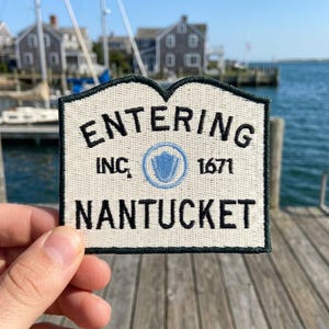 May include: A rectangular embroidered patch with a cream-colored background and a dark green border. The text reads ENTERING, INC. 1671, and NANTUCKET in black, with a central light blue circular emblem featuring a shield icon inside the text.