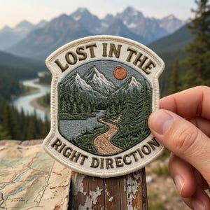 May include: A circular embroidered patch featuring a mountain landscape with a winding path, a river, and a sun. The text "LOST IN THE RIGHT DIRECTION" is stitched in dark brown thread along the top and bottom edges of the cream-colored border.