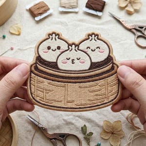 May include: An embroidered patch featuring three cream-colored bao buns with smiling faces and pink cheeks, nestled inside a tan bamboo steamer basket. The design uses brown thread for the basket details and outlines, creating a textured appearance.