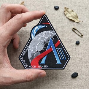 May include: A triangular embroidered patch with a black background featuring a gray moon, a blue Earth, a red curved line, and a large blue letter A with two vertical lines. The text reads WISEMAN, GLOVER, and KOCH HANSEN in white capital letters.
