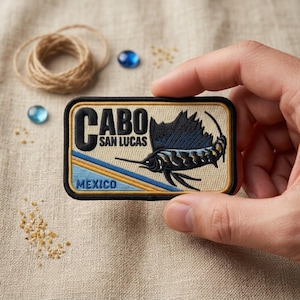 May include: A rectangular embroidered patch featuring a black sailfish graphic, the text "CABO SAN LUCAS" in black, and "MEXICO" in blue. The design includes a beige background with diagonal blue and yellow stripes, all bordered by a black edge.