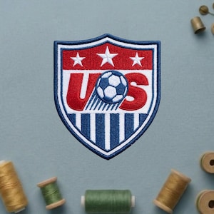 May include: An embroidered shield-shaped patch featuring a red, white, and blue design. The top section is red with three white stars. The center displays the letters US in red with a soccer ball replacing the letter S. The bottom has blue stripes.
