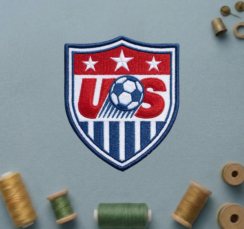May include: An embroidered shield-shaped patch featuring a red, white, and blue design. The top section is red with three white stars. The center displays the letters US in red with a soccer ball replacing the letter S. The bottom has blue stripes.
