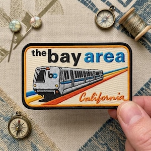 May include: A rectangular embroidered patch featuring a white train with blue stripes and the text "the bay area" in black and blue, with "California" in orange script. The design includes diagonal orange, yellow, and blue stripes.