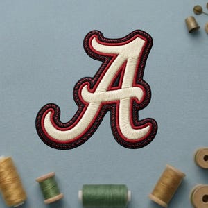 May include: Embroidered letter "A" patch in cream, red, and black. The letter has a rope-like border and is set against a light blue background. Sewing notions, including spools of thread, are scattered around the patch.