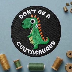 May include: Embroidered patch with a green dinosaur illustration on a black circular background. The text "DON'T BE A CUNTASAURUS" is embroidered in white around the dinosaur. The patch is surrounded by spools of thread.
