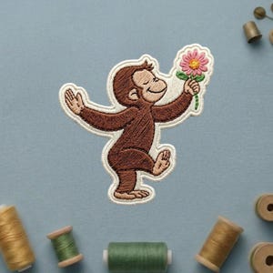 May include: An embroidered patch featuring a brown monkey with a smiling expression, holding a pink flower with a yellow center. The patch has a white border and is rendered in a cartoon style with detailed stitching throughout the design.