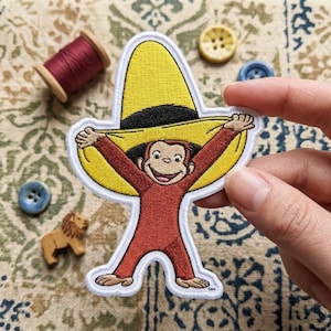 May include: An embroidered patch featuring a brown monkey wearing a large yellow sombrero with a black band. The monkey has a smiling expression and is depicted with arms raised, holding the brim of the hat. The patch has a white border.