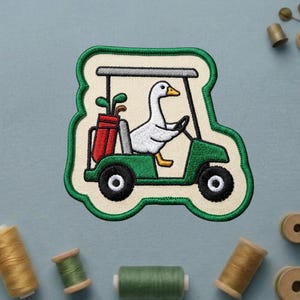 May include: Embroidered patch featuring a white goose driving a green golf cart. The golf cart has black wheels and a red golf bag with clubs. The patch has a cream-colored background and a green border. A fun, whimsical design for golf enthusiasts.