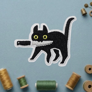 May include: Embroidered black cat patch with yellow eyes and a knife in its mouth. The cat has a white outline and is on a light blue background. Sewing notions, including spools of thread and pins, are scattered around the patch.