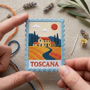 May include: A rectangular embroidered patch with a scalloped light blue border featuring a landscape of a house in Tuscany. The scene includes a yellow house with a red roof, cypress trees, rolling fields, a blue path, and the text TOSCANA.