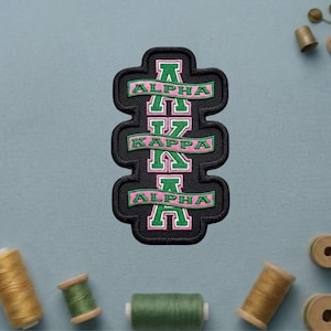 May include: Embroidered patch featuring the Greek letters Alpha Kappa Alpha in green and pink on a black background. The patch has a unique, layered design and is surrounded by a green border. The patch is likely for a sorority.