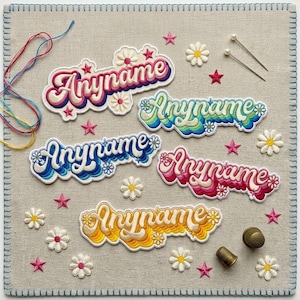 May include: Five embroidered patches featuring the text Anyname in various color gradients including pink, blue, green, and yellow. The patches are decorated with small embroidered daisies and stars, accompanied by sewing needles and thimbles.