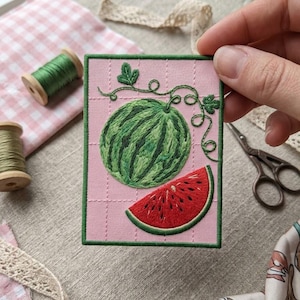 May include: A rectangular embroidered patch featuring a light pink background with a grid pattern. The design displays a whole green striped watermelon and a single red watermelon slice with black seeds, accented by green embroidered vines.
