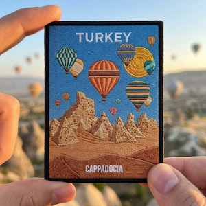 May include: A rectangular embroidered patch featuring a blue sky with multiple colorful hot air balloons and a yellow sun above tan rock formations. The text TURKEY is at the top and CAPPADOCIA is at the bottom in white capital letters.