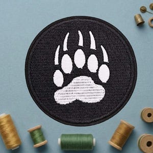 May include: A round, black embroidered patch featuring a white bear paw print. The patch is surrounded by a black border. Sewing supplies, including spools of thread and a thimble, are arranged around the patch.