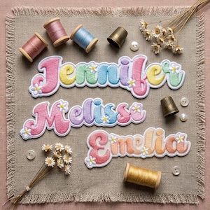 May include: Three embroidered name patches featuring the names Jennifer, Melissa, and Emelia in a colorful, rounded font with small white flower accents. The patches are arranged on a beige fabric square with spools of thread and thimbles.