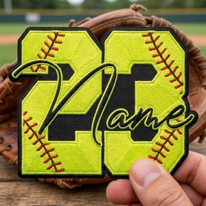 May include: A neon yellow embroidered patch in the shape of the number 23, featuring black outlines and red stitching details that mimic a softball. The word Name is written across the center in a black cursive font with a neon yellow border.